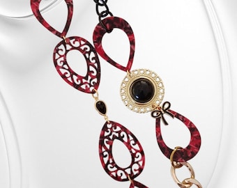 May include: A striking necklace with a combination of elements. It features a black chain, red and black teardrop pendants, and a gold-toned chain. A gold-toned circular pendant with a black stone and a small bow detail adds a touch of sophistication.