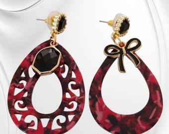 May include: A pair of teardrop earrings with a red and black marbled pattern. One earring features a black gemstone and gold-coloured accents, while the other has a black bow. The earrings have gold-coloured posts and black accents.