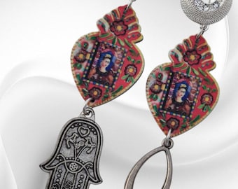 May include: Dangle earrings with silver-coloured metal details. Each earring has a red heart-shaped pendant with a portrait and floral design, and a silver charm. One earring has a Hamsa hand charm, the other a wishbone.
