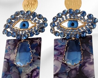 May include: A pair of statement earrings featuring an eye design. Each earring has a gold-colored disc, an eye-shaped design with blue crystals, and a rectangular pendant with a blue and purple marbled pattern.