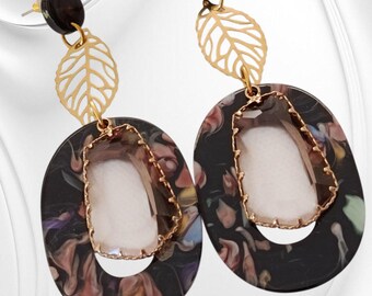 May include: A pair of dangle earrings. Each earring has a black oval with a floral pattern, a gold leaf, and a clear, rectangular stone. The earrings have gold-coloured metal accents and a black stud.