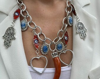May include: A silver chain necklace with multiple charms, including a red evil eye, a blue and white eye, a silver hand, a red horn, and a silver heart.