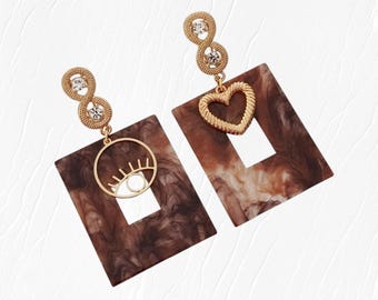 May include: A pair of gold-toned earrings. Each earring features a rectangular brown and white marbled design with a cut-out square. One earring has a gold eye design, the other a gold heart. The earrings have a gold chain with three decorative circles.