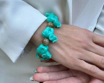 May include: A turquoise and brown beaded bracelet and necklace set. The bracelet has irregular turquoise beads and round brown beads. The jewellery is worn on a wrist, with a white blazer in the background.