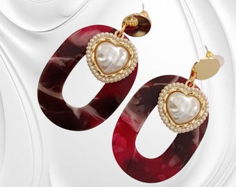 May include: A pair of dangle earrings. Each earring features a maroon and white oval shape with a gold-coloured heart pendant. The heart pendant is adorned with a pearl-like centre and a border of small pearls. The earrings have gold-coloured posts.
