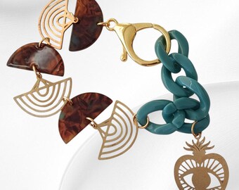 May include: A gold-toned bracelet with a teal chain and a gold clasp. The bracelet features brown and gold geometric shapes and a gold heart-shaped charm with an eye design. The bracelet is on a white background.