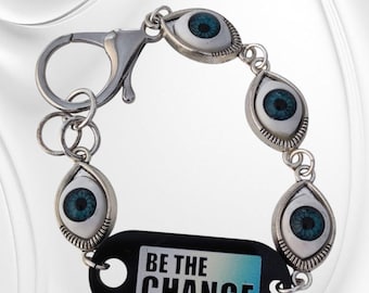 May include: Silver bracelet with five eye-shaped charms, each with a blue iris and black pupil. A black tag with the words "BE THE CHANGE" is attached. The bracelet has a clasp closure.