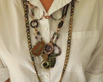 May include: A multi-layered necklace featuring a range of charms, including a green camouflage chain, a copper chain, a black chain, and a clear plastic chain with a green camouflage pattern. The charms include a round eye charm, a grenade charm, a "BOOM" charm, and a clear plastic charm with a green camouflage pattern.