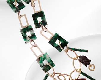 May include: A statement necklace featuring a mix of geometric shapes and charms. The necklace has gold-tone chain links, green and white marbled squares, and rectangular accents. Charms include a Buddha head, a heart, and an eye pendant.