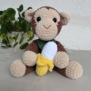 Op de afbeelding: Een pluche apenknuffel gemaakt van bruin en beige garen met zwarte plastic ogen. De aap heeft een donkergroene kraag en houdt een geel met witte gehaakte banaan vast. Het speelgoed heeft een zachte, getextureerde en pluizige garenstructuur.