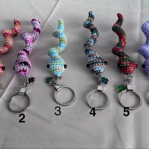 Op de afbeelding: Een verzameling van zes handgemaakte gehaakte sleutelhangers. Elke sleutelhanger heeft een uniek, kleurrijk, spiraalvormig ontwerp met een kleine, ronde kop en een metalen sleutelring. De sleutelhangers zijn in verschillende kleuren, waaronder roze, paars, blauw en groen.