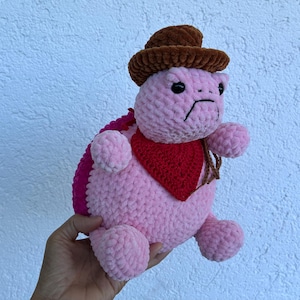 May include: A plush, round, pink crocheted turtle toy featuring a brown textured cowboy hat, a red triangular bandana around its neck, and black bead eyes. The turtle has a bright pink shell on its back and a soft, fuzzy yarn texture throughout.