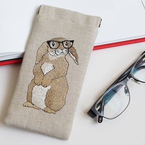 Può includere: Un astuccio per occhiali in lino beige con un coniglio marrone ricamato che indossa gli occhiali. Il coniglio è in piedi con le zampe unite.