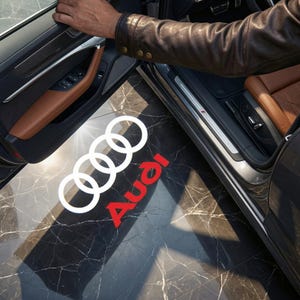 Puede incluir: El logotipo de Audi, que consta de cuatro anillos blancos entrelazados y la palabra "Audi" en rojo, se proyecta sobre un suelo de mármol junto a una puerta de coche abierta. El interior del coche presenta asientos de cuero marrón y un panel de puerta oscuro.