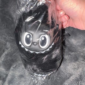 Könnte beinhalten: Ein schwarzes Plüschtier mit großen, weiß umrandeten Augen und einem weißen, gezackten Grinsen, verpackt in durchsichtigem Kunststoff. Das Spielzeug hat eine flauschige Textur und wird in einer Hand vor grauem Hintergrund gehalten.