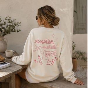 Könnte beinhalten: Ein cremefarbenes Langarm-Sweatshirt mit roter Strichzeichnung auf der Rückseite. Das Design zeigt den Schriftzug "nurse michelle" in Schreibschrift, eine Schleife, eine Spritze, ein Kasack-Oberteil, ein Pflaster, ein Stethoskop und Sterne.