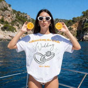 Könnte beinhalten: Ein weißes T-Shirt mit dem orangefarbenen Text Vollstreckung des Urteils und JGA Jacky sowie einer schwarzen Strichzeichnung von zwei ineinander verschlungenen Ringen, einer davon mit Diamant, umgeben von kleinen Sternen, getragen mit Orangenscheiben.