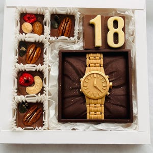 Könnte beinhalten: Eine weiße Schachtel mit Pralinen. Die Schachtel enthält Pralinen mit Nüssen und Trockenfrüchten, eine Schokoladen-Uhr und einen Schokoladenblock mit der Zahl 18 in Gold. Die Pralinen sind auf weißem Papier angeordnet.