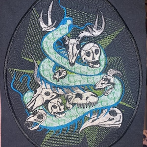 Puede incluir: Parche bordado ovalado con un diseño de serpiente turquesa entrelazada con calaveras de animales. El fondo negro presenta un patrón geométrico verde neón. El diseño es detallado e intrincado.