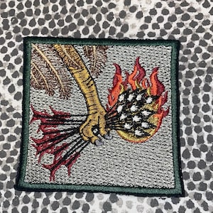May include: Embroidered patch featuring a golden claw clutching a bundle of arrows engulfed in flames. The patch has a green border and a textured gray background. The flames are red and orange, and the arrows are black and white.