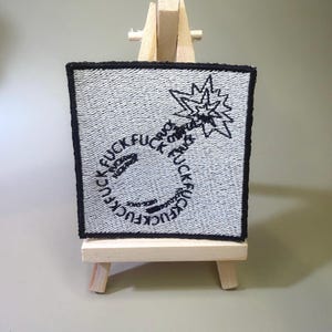 May include: A square embroidered patch with a white background and a black border. The design features a bomb shape outlined by the repeated word "FUCK" in black thread, with a small explosion graphic at the top and additional text inside.