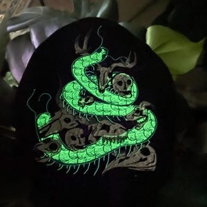 K&ouml;nnte beinhalten: Schwarze M&uuml;tze mit einem leuchtend gr&uuml;nen Schlangenmuster, das sich um Sch&auml;del und Geweihe windet. Das Design hat ein detailliertes, strukturiertes Aussehen, wobei die Schuppen der Schlange und die Merkmale der Sch&auml;del deutlich sichtbar sind. Das Gesamtthema ist gotisch und makaber.
