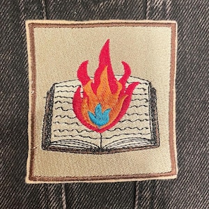 May include: Embroidered patch featuring an open book with flames rising from the center. The flames are red, orange, and blue, set against a beige background with a brown border. The patch is likely for clothing or accessories.