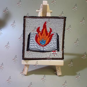 May include: A square embroidered patch featuring an open book with a large orange and red flame rising from the center, accented by a small blue flame at the base. The patch has a light gray background, a dark brown border, and the text Darned Anarchist.