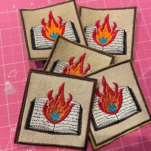 K&ouml;nnte beinhalten: Gestickte Aufn&auml;her mit einem offenen Buch mit Flammen. Die Aufn&auml;her haben einen beigefarbenen Hintergrund mit einem braunen Rand und ein farbenfrohes Flammendesign in Rot, Orange und Blau. Die Aufn&auml;her sind quadratisch.