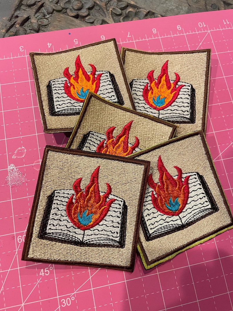 K&ouml;nnte beinhalten: Gestickte Aufn&auml;her mit einem offenen Buch mit Flammen. Die Aufn&auml;her haben einen beigefarbenen Hintergrund mit einem braunen Rand und ein farbenfrohes Flammendesign in Rot, Orange und Blau. Die Aufn&auml;her sind quadratisch.