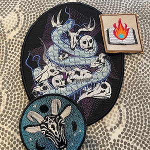 May include: Embroidered patches featuring occult and gothic designs. Includes a large oval patch with a snake coiled around skulls, a square patch with a burning book, and a round patch with a goat head and pentagram.