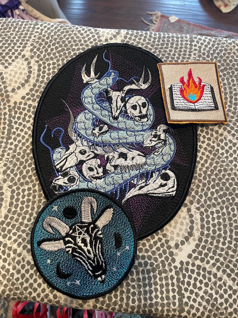 K&ouml;nnte beinhalten: Gestickte Aufn&auml;her mit gotischen und okkulten Designs. Das Bild zeigt einen ovalen Aufn&auml;her mit einer Schlange und Totenk&ouml;pfen, einen quadratischen Aufn&auml;her mit einem brennenden Buch und einen runden Aufn&auml;her mit einem Ziegenkopf und einem Pentagramm.