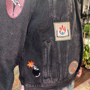 K&ouml;nnte beinhalten: Schwarze Jeansjacke mit aufgestickten Aufn&auml;hern. Aufn&auml;her zeigen ein brennendes Buch, eine Bombe und ein Friedenszeichen. Ein Aufn&auml;her lautet "SO FUN IT HURTS". Die Jacke hat zwei Vordertaschen und einen Knopfverschluss.