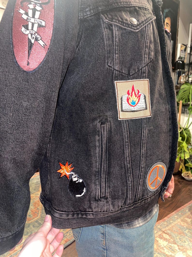K&ouml;nnte beinhalten: Schwarze Jeansjacke mit aufgestickten Aufn&auml;hern. Aufn&auml;her zeigen ein brennendes Buch, eine Bombe und ein Friedenszeichen. Ein Aufn&auml;her lautet "SO FUN IT HURTS". Die Jacke hat zwei Vordertaschen und einen Knopfverschluss.