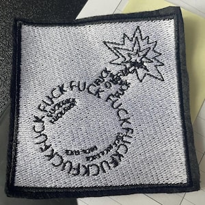 May include: A square embroidered patch with a white background and a black border. The design features a bomb shape outlined in black thread, with the word "FUCK" repeated multiple times to form the bomb's body and a starburst at the top.