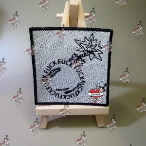 May include: A square embroidered patch with a white background and a black border. The design features a black outline of a bomb with a fuse, surrounded by the repeated word FUCK in a circular pattern. A small heart with a dagger and the text Darned Anarchist is at the bottom right.