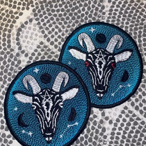 May include: Two circular embroidered patches. Each patch features a goat's head with a pentagram on a teal background. The goat has white horns and a black and white face with red eyes. The patches have a black border and star and moon details.