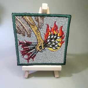 May include: A square embroidered patch featuring a yellow bird claw holding a bundle of thin black sticks that are ignited in bright orange and red flames. The background is a light gray textured stitch pattern with a dark green border edge.