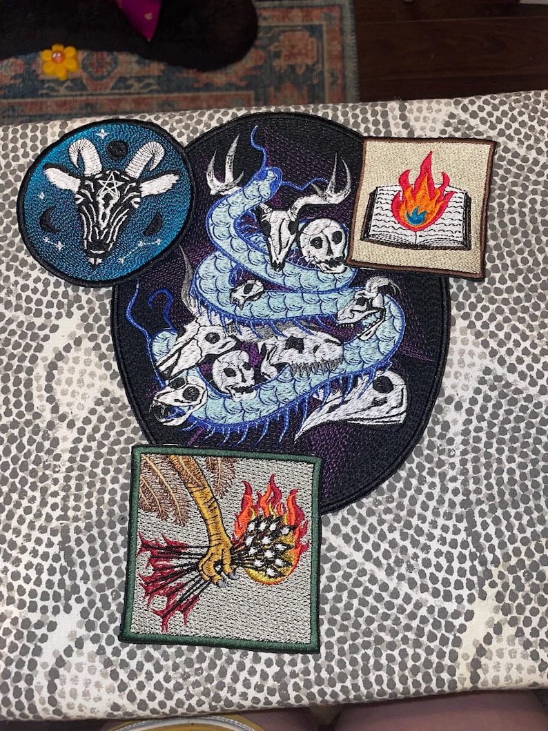 May include: Embroidered patches featuring occult and gothic designs. Includes a blue circular patch with a goat head, a large patch with a snake and skulls, and two square patches with flames and a hand holding arrows.