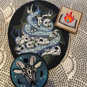 K&ouml;nnte beinhalten: Gestickte Aufn&auml;her mit gotischen und okkulten Designs. Das Bild zeigt einen gro&szlig;en schwarzen Aufn&auml;her mit einem Schlangen- und Totenkopfmotiv, einen quadratischen Aufn&auml;her mit einem brennenden Buch und einen runden Aufn&auml;her mit einem Ziegenkopf und einem Pentagramm.