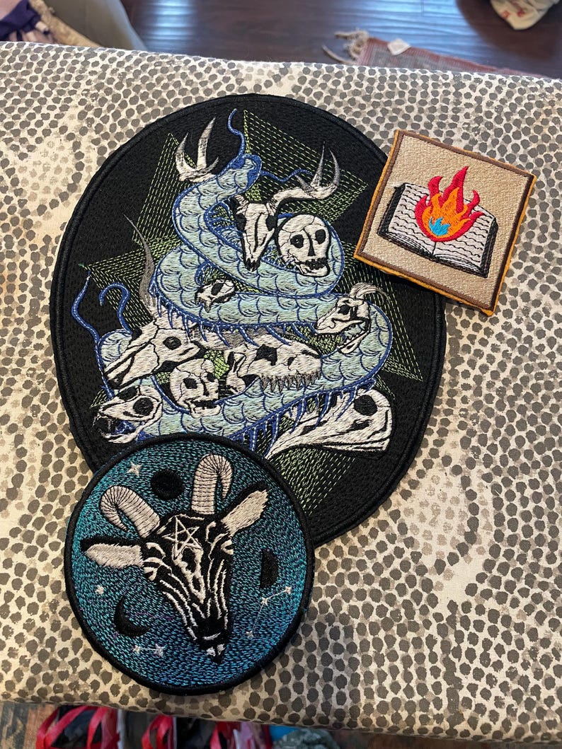K&ouml;nnte beinhalten: Gestickte Aufn&auml;her mit gotischen und okkulten Designs. Das Bild zeigt einen gro&szlig;en schwarzen Aufn&auml;her mit einem Schlangen- und Totenkopfmotiv, einen quadratischen Aufn&auml;her mit einem brennenden Buch und einen runden Aufn&auml;her mit einem Ziegenkopf und einem Pentagramm.