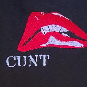 May include: Black textile with an embroidered design featuring a pair of red lips with white teeth and the word "CUNT" in white capital letters below the lips.