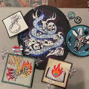 May include: A collection of embroidered patches including a large black oval patch with a blue snake and skulls, a square patch with a burning book, a square patch with a hand holding arrows in fire, a square patch with text, and a circular goat patch.