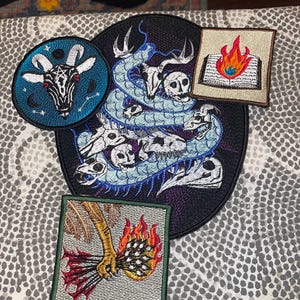 May include: A collection of embroidered patches featuring gothic and occult designs. Includes a round patch with a goat head, a large patch with a snake and skulls, and two square patches with a burning book and a hand holding arrows.