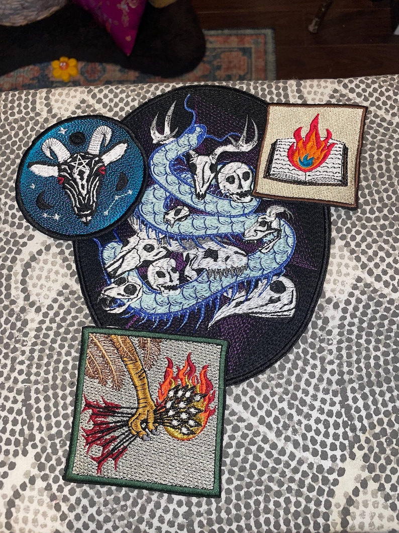 May include: A collection of embroidered patches featuring gothic and occult designs. Includes a round patch with a goat head, a large patch with a snake and skulls, and two square patches with a burning book and a hand holding arrows.