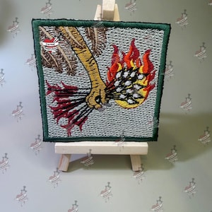 May include: A square embroidered patch with a dark green border featuring a yellow hand holding a bundle of black arrows that are engulfed in bright red and orange flames against a textured light gray background with a small logo in the corner.