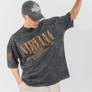 Könnte beinhalten: Ein anthrazitfarbenes T-Shirt in Acid-Wash-Optik mit dem Text NIRVANA, THE BUZZCOCKS, MIERCOLES 9 de FEBRERO und PALACIO DE LOS DEPORTES in beiger Schrift, kombiniert mit einer passenden Baseballkappe in Acid-Wash-Optik mit New York Authentic.
