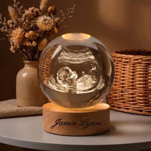 May include: A clear glass sphere with an illuminated ultrasound image of a baby, resting on a wooden base. The base is engraved with the name "Jamie Lynn". The sphere is lit from within, creating a warm glow. A vase of dried flowers and a wicker basket are in the background.