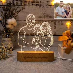 May include: A family portrait night light with a wooden base. The light features a line drawing of a family of three, illuminated with a warm glow. The base is engraved with the words "My family, my everything."
