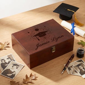 May include: A rectangular dark brown wooden keepsake box with a metal latch, featuring a laser-engraved graduation cap design and the name Jamie Lynn. The box is surrounded by a black graduation cap, a rolled diploma, a fountain pen, and photos.